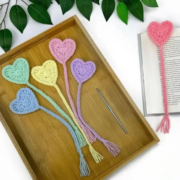 Five crochet heart bookmarks are displayed on a wooden tray, with an open book and a sixth bookmark placed inside the book.