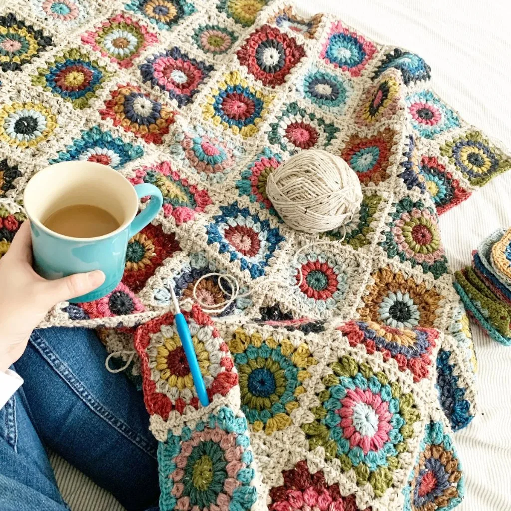 A hand holding a coffee cup over a cozy crochet blanket made from colorful yarn scraps, showcasing a warm and creative stash-busting projec