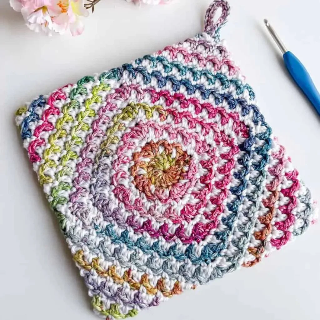 A colorful crochet potholder with a hanging loop with a crochet hook and flowers.