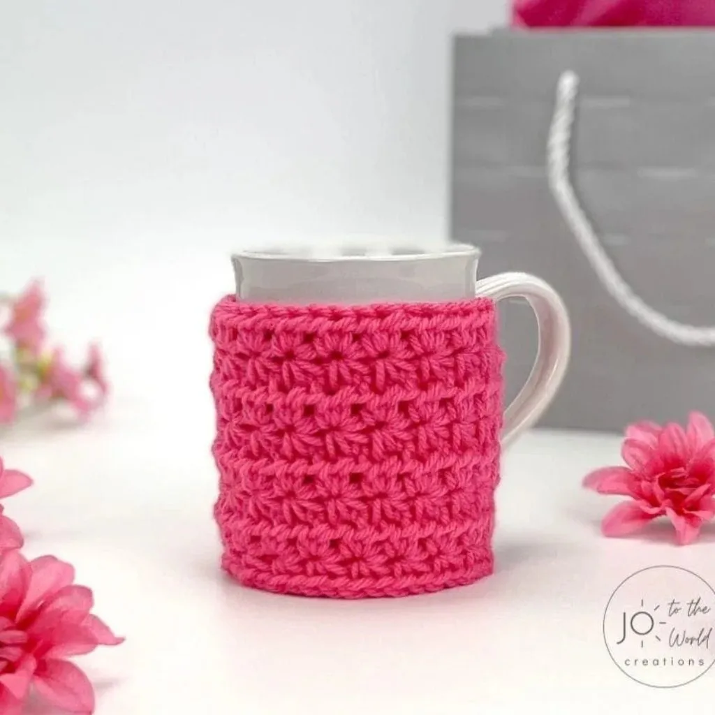 A coffee cup in a crochet sleeve with a gift box and flowers.