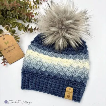 Blue ombré crochet beanie with ribbed brim and puff stitch texture, finished with a faux fur pom pom — free crochet hat pattern for women