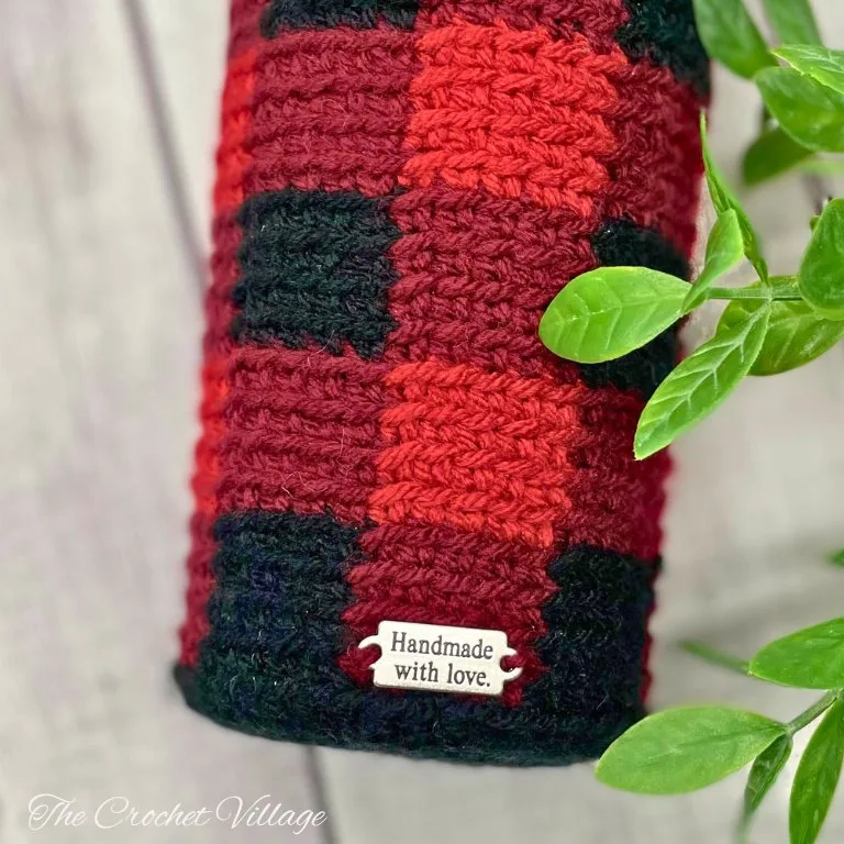 A crochet piece featuring a buffalo check plaid pattern in red, brown, and black colors, adorned with a metal tag.