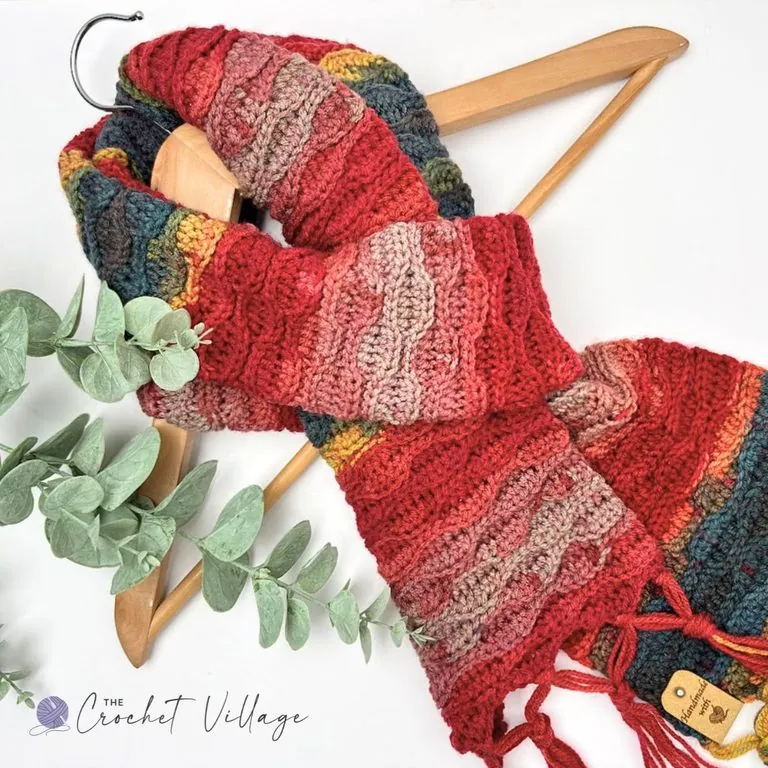 Tamara Scarf free crochet pattern in red, teal, and gold variegated yarn with wave stitch texture and fringe, draped on a wooden hanger