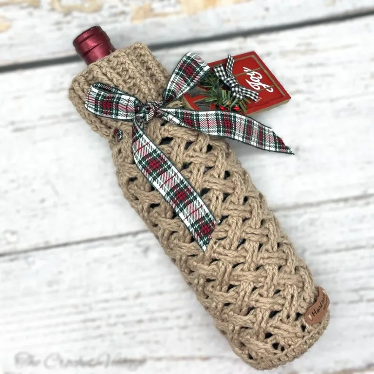 A wine bottle wrapped in a festive crochet Celtic weave stitch cozy, decorated with a Christmas tag for the perfect holiday gift.