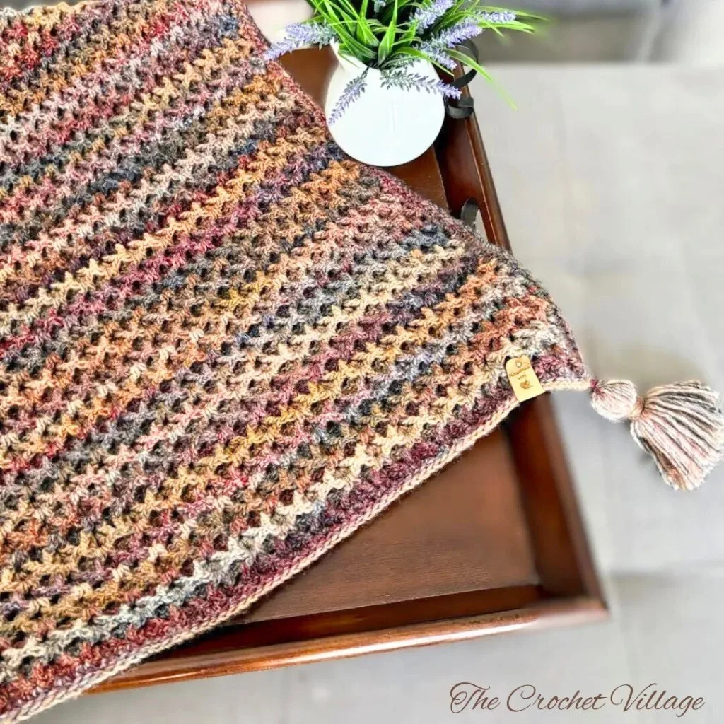 A crochet piece with a tassel featuring the waffle V stitch on a wooden surface, accompanied by a vase and flowers.