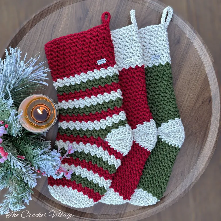 Three textured, large festive crochet Christmas stockings, including one striped design, adorned with holiday picks for a cozy seasonal display.