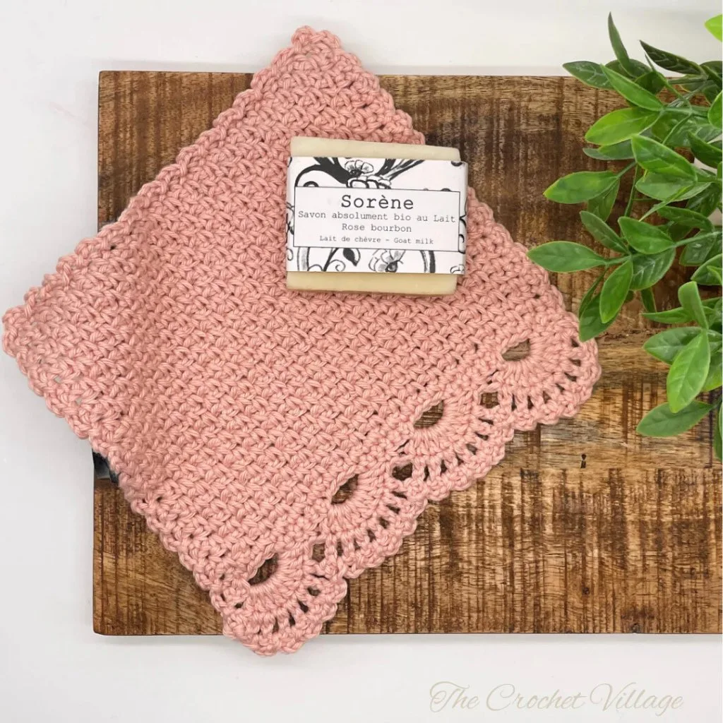 A fancy crochet washcloth with a delicate lacy border, elegantly displayed with a bar of soap and a small plant.