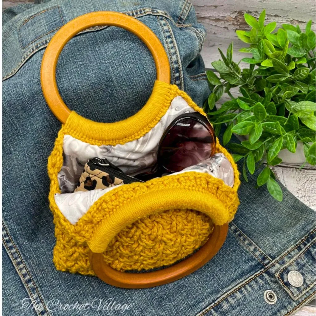 An open crochet handbag with wooden handles, lined with fabric, carrying a wallet and glasses. The bag rests on a jean jacket.