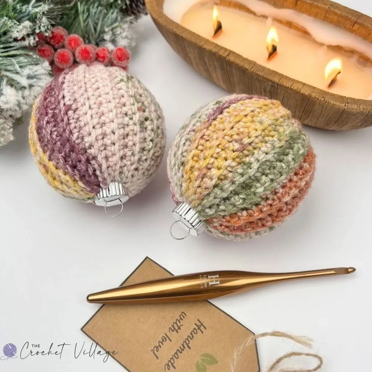 Two crochet Christmas bauble covers in variegated pink, green, and gold yarn beside a gold crochet hook and a handmade gift tag