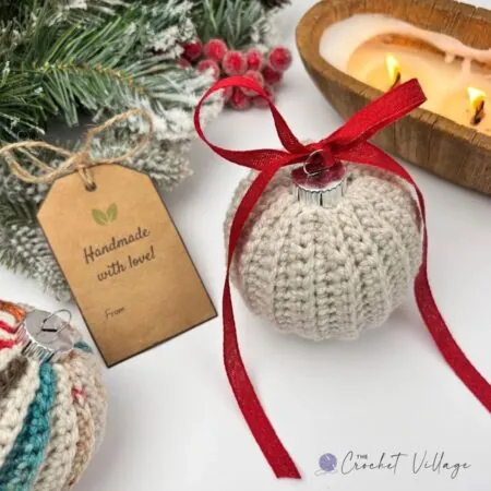 Cream crochet Christmas bauble cover tied with a red ribbon bow, beside a handmade gift tag and a lit candle