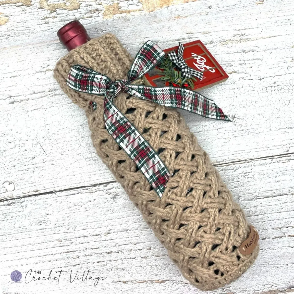 Tan crochet wine bottle cozy with Celtic Weave stitch, tied with a plaid Christmas ribbon and gift tag