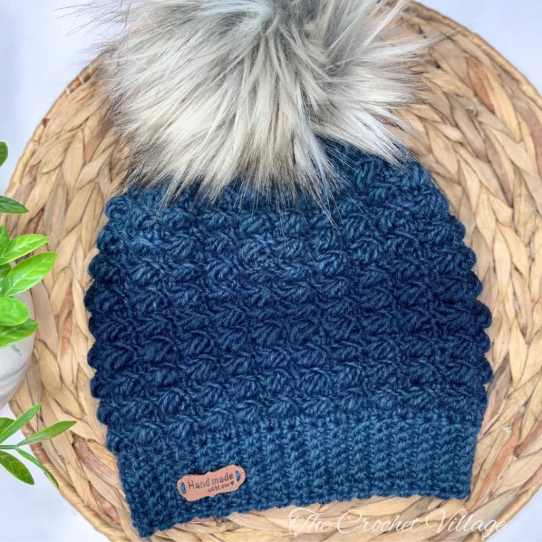 A textured crochet hat with a large pom pom on a wooden surface