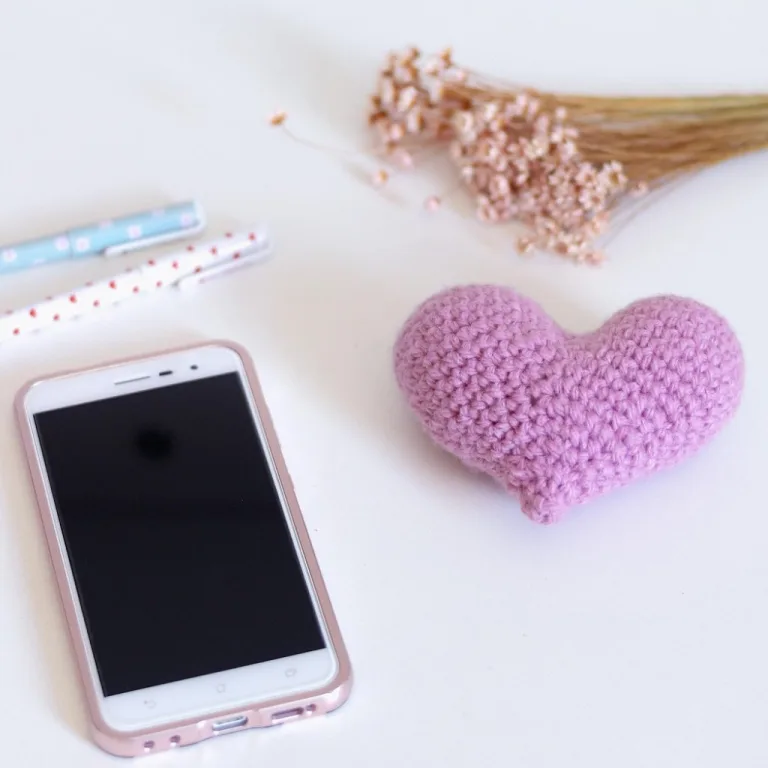 A crocheted heart placed beside a smartphone, two pens, and a small plant, set up for crochet photography.