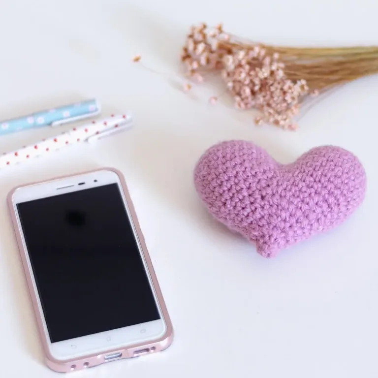 A crocheted heart placed beside a smartphone, two pens, and a small plant, set up for crochet photography.