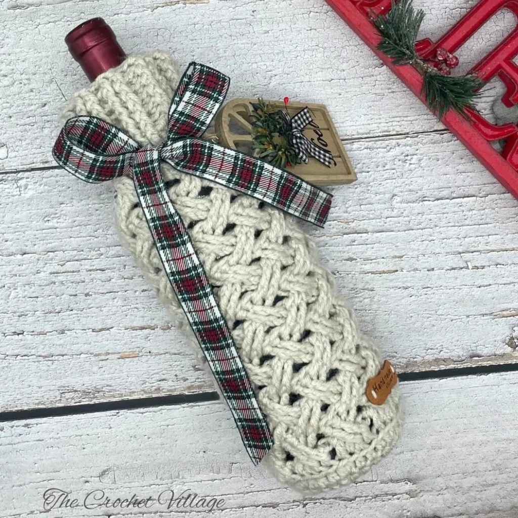 A wine bottle wrapped in a festive crochet Celtic weave stitch cover, decorated with a Christmas tag for the perfect holiday gift.