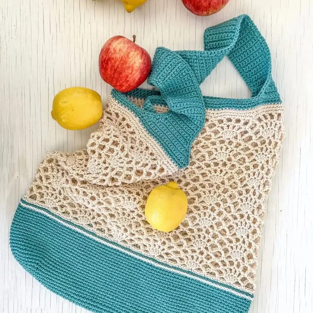A crochet market tote bag featuring a lacy stitch design, a sturdy base, and durable handles, filled with oranges and apples.