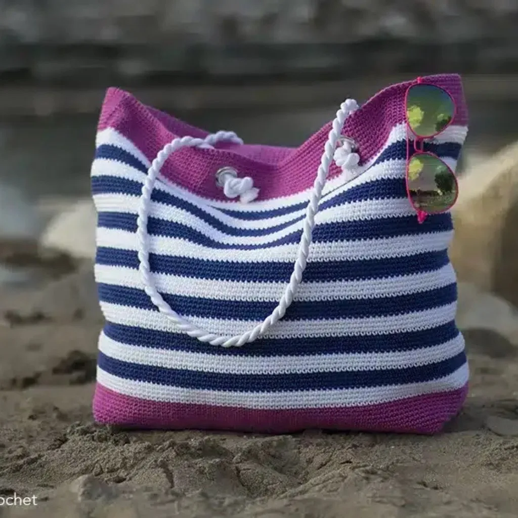 A crochet beach bag with a nautical design, sturdy cord handles, and sunglasses resting on the sand.