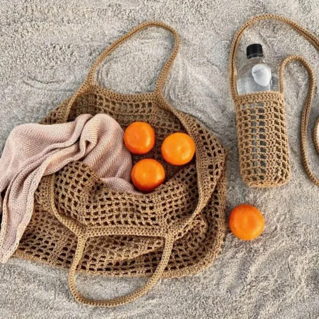 A crochet mesh tote filled with a few oranges, resting on the sand, alongside a bottle of water in a matching crochet mesh bottle cozy.