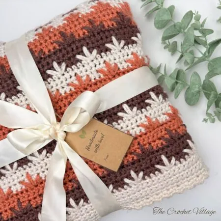 Cozy crochet afghan in warm fall colors, draped over a chair, featuring autumn leaves in rich shades of orange, brown, and beige