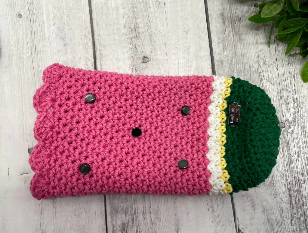 A crochet watermelon-themed wine bottle sleeve with small black buttons sewn on to represent seeds.