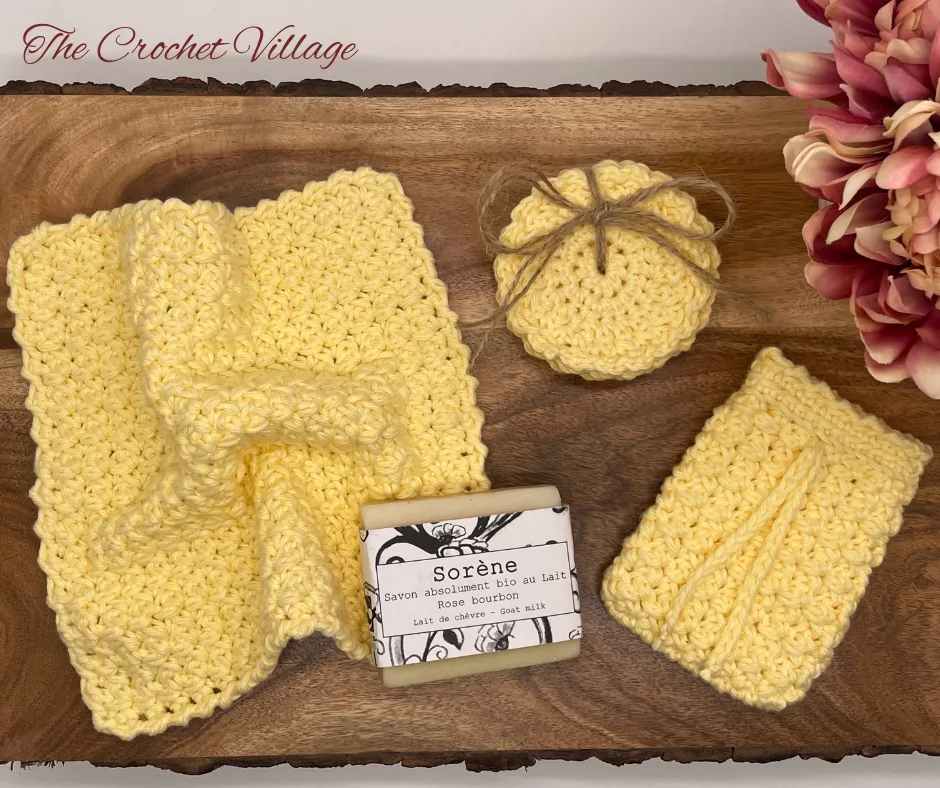 A handmade, eco-friendly crochet bathroom set displayed on a wooden surface, including a reusable washcloth, a soap saver pouch, and round face scrubbies, shown with a bar of soap.