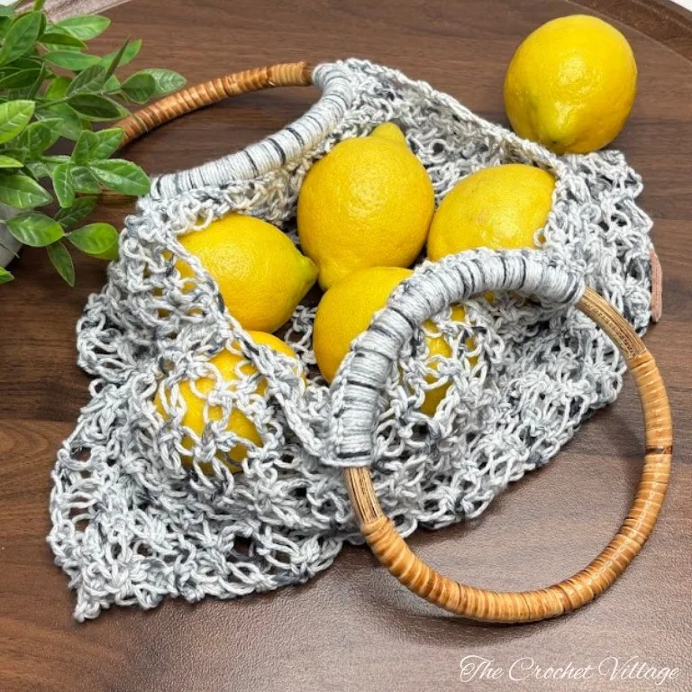 Crochet produce bag made with a lacy Solomon’s Knot stitch, featuring round wooden handles. The bag is filled with fresh yellow lemons and placed on a rustic wooden surface.