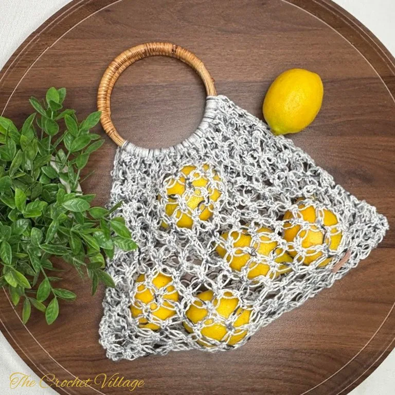 Crochet market bag made with a lacy Solomon’s Knot stitch, featuring round wooden handles. The bag is filled with fresh yellow lemons and placed on a rustic wooden surface.