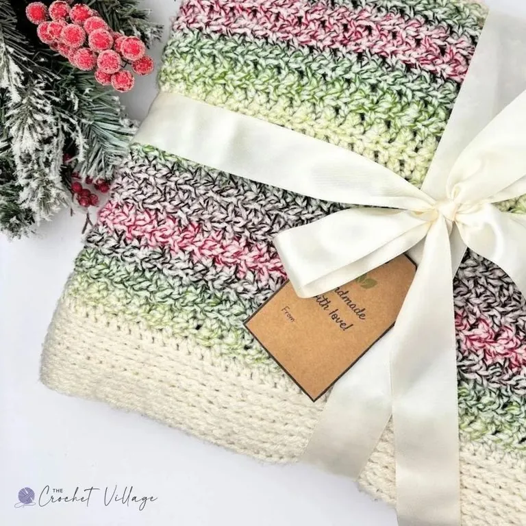 Winterland crochet blanket folded and tied with a cream satin bow and handmade gift tag beside frosted pine and red berries