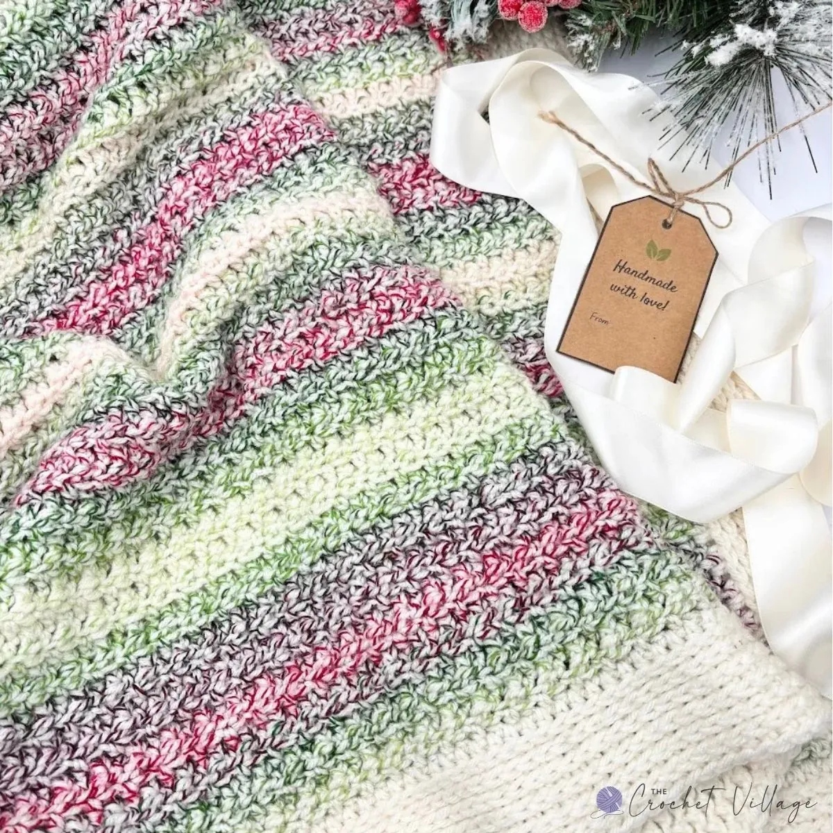 Winterland Blanket free crochet pattern in variegated pink, green, and cream stripes with a cream ribbon and handmade gift tag