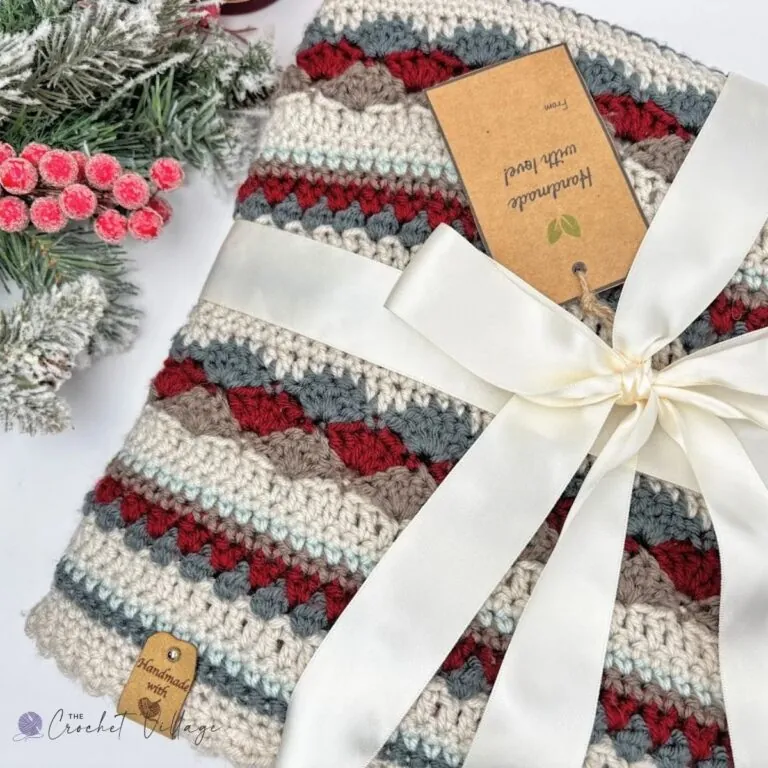 Heartland scrap yarn crochet blanket folded with a cream ribbon and handmade gift tag beside frosted pine and red berries