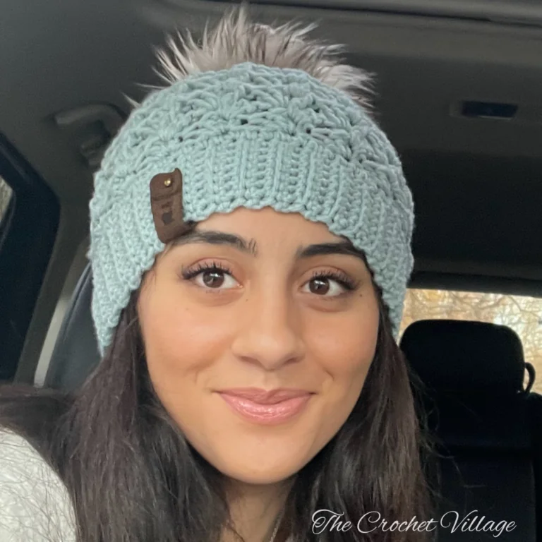 Front view of a woman wearing the light blue Alpine Hat crochet beanie, showing the ribbed brim fit and grey faux fur pom-pom