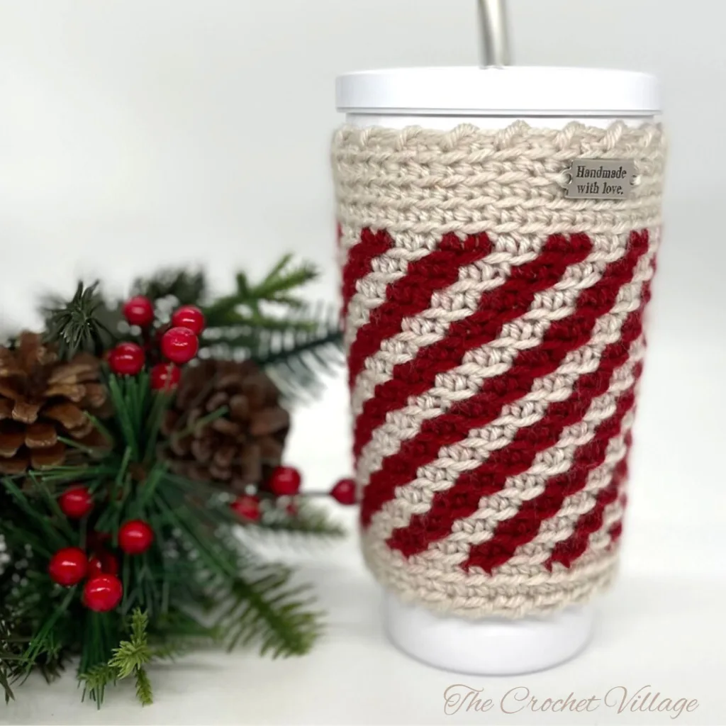 White cup in a handmade crochet striped peppermint cozy, surrounded by festive Christmas decor and holiday ornaments.