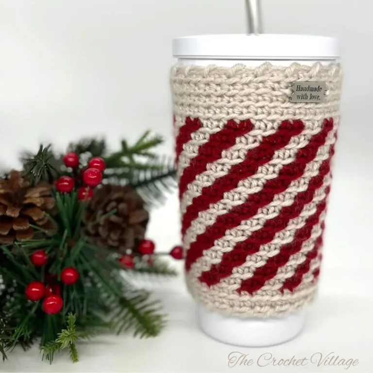 White cup in a handmade crochet striped peppermint cozy, surrounded by festive Christmas decor and holiday ornaments.