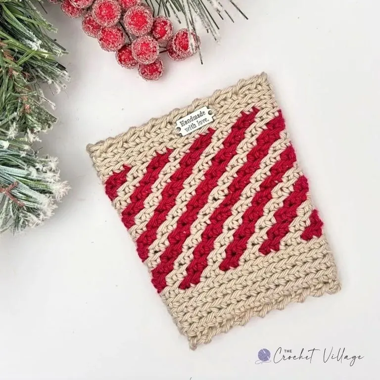 Crocheted cup cozy featuring a festive red and beige striped design with a 'Handmade with Love' tag, perfect for adding a cheerful touch to holiday beverages.