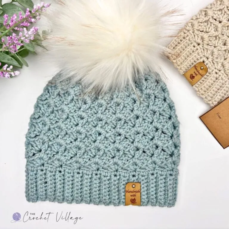 Light blue Alpine Hat free crochet pattern with ribbed brim, textured stitch, and cream faux fur pom-pom, styled with pink flowers and a gift tag