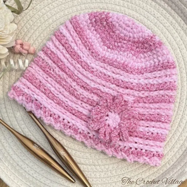 A pink variegated crochet chemo hat made from top down with two crochet hooks and a lit candle beside it