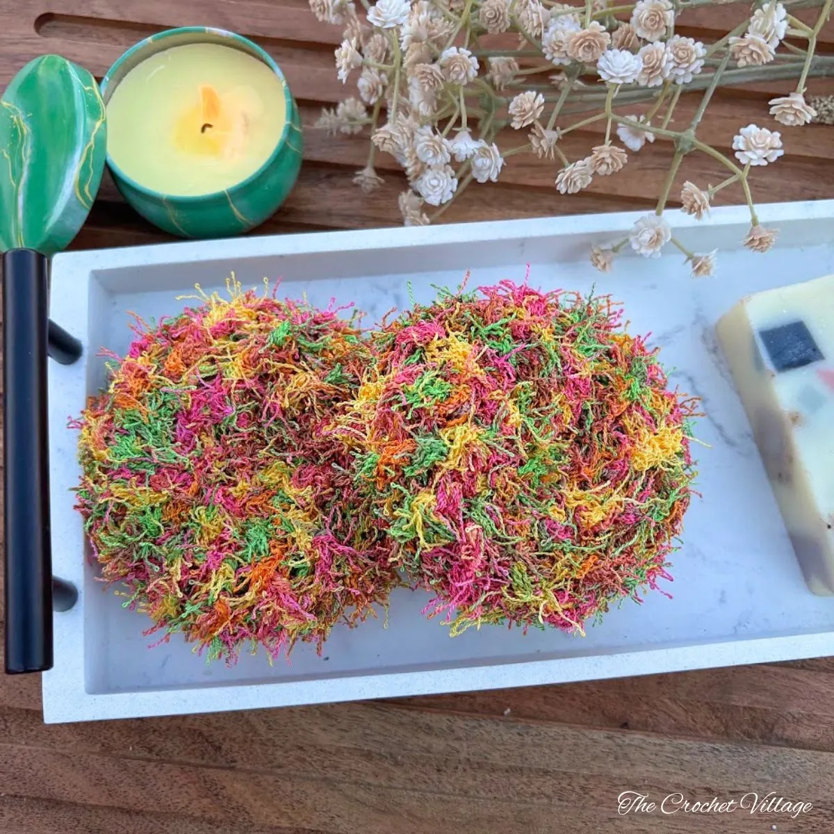 Two crochet scrubbies in multicolor yarn on a marble tray with candle and soap