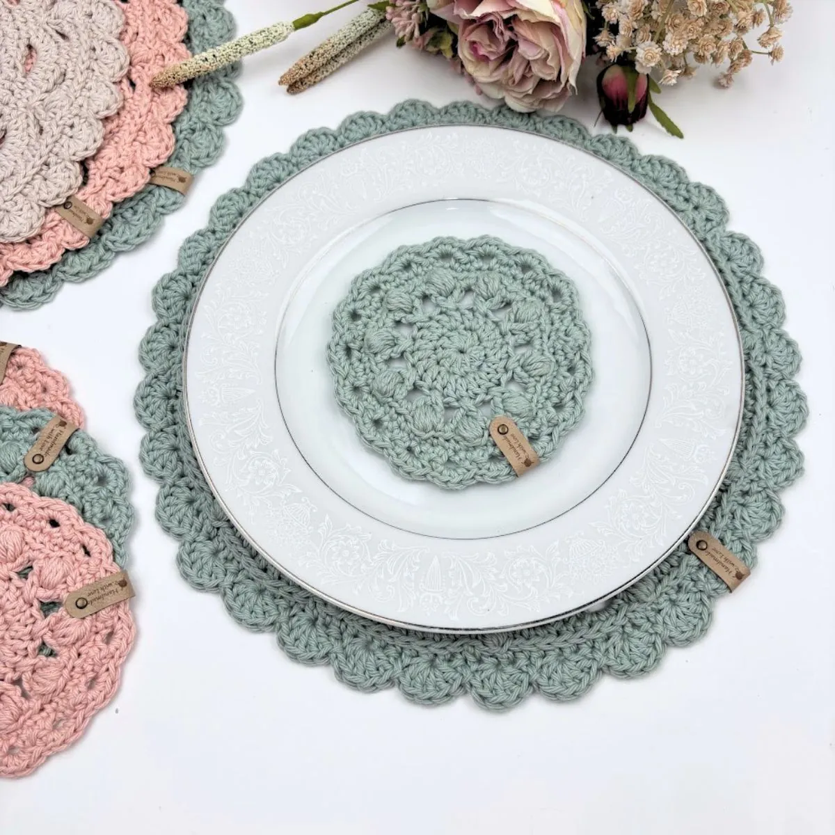 Sage green round crochet placemat with scalloped edge under a white floral plate, with a matching crochet coaster