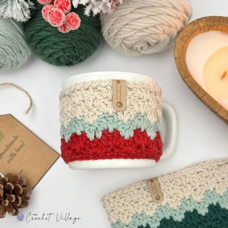 Crochet mug cozy in cream, sage, and red on a white mug, with a second cozy in green and cream lying flat