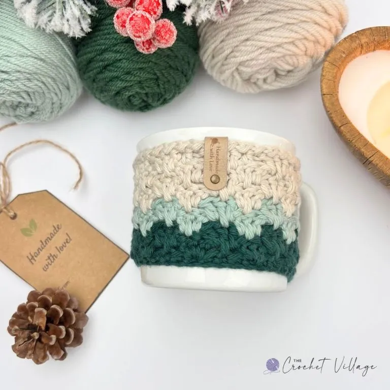 Crochet mug cozy in forest green, sage, and cream on a white mug with a handmade leather tag